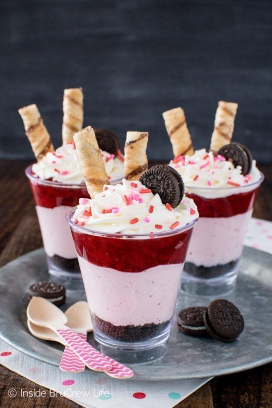 Three small transparent cups sit on a silver plate. Each cup is filled with a bottom-to-top layer of crushed oreos, strawberry cheesecake, and whipped cream with red and pink sprinkles. The parfaits are topped with one Oreo cookie and one pipe-like wafer cookie. The plate also has three Oreos and a pile of three wooden dessert spoons on it.