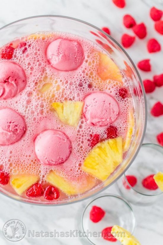 A top-down view of light pink punch in a large glass bowl on a marble countertop. The punch’s surface is covered in bubbles. Pink scoops of ice cream, raspberries, and chunks of pineapple float in the punch. There are two empty glasses and a dozen raspberries on the counter to the right of the bowl.