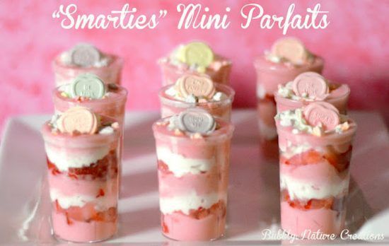 Nine Smarties mini parfaits are arranged in a three-by-three grid on a white square platter. Each parfait is in a transparent cylindrical cup and layered from bottom-to-top with pink yogurt-and-Smarties mixture, then strawberries, and finally whipped cream. The layering repeats twice, and each parfait is garnished with a Smarties candy.